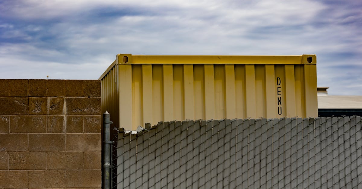 Yellow shipping container behind industrial fence under cloudy sky in urban setting.